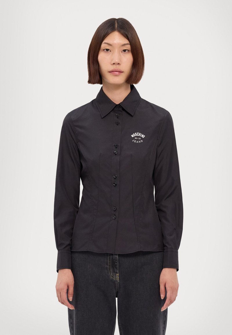 Donna con capelli castani lisci indossa una camicia nera attillata con il logo Moschino Jeans sul petto e jeans scuri, in piedi contro uno sfondo bianco.
