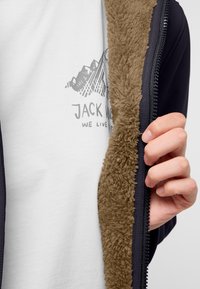 Fleecegefütterte Zip-Jacke mit einer dunklen Außenschale und einem weichen, tan-farbenen Innenfutter. Besitzt einen grafischen Druck auf einem weißen T-Shirt darunter.