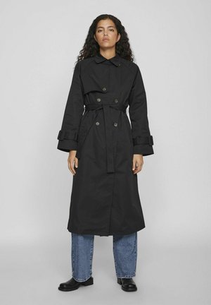 Cappotto lungo nero con vita alta e cintura, bottoni doppi e maniche larghe. Indossato sopra jeans blu chiaro e stivali neri.