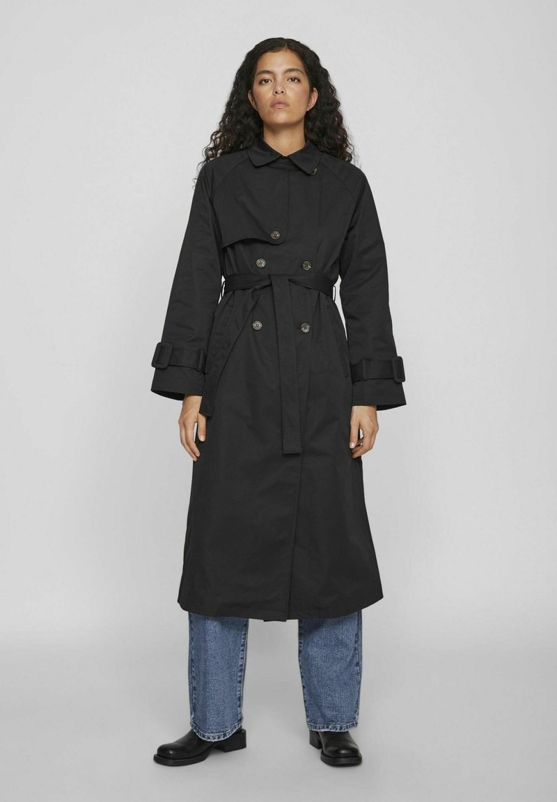 Melnā tranša mētelis ar jostu viduklī, dubulto pogu aizdari un plašām piedurknēm. Valkāts pār gaiši ziliem džinsiem un melnām zābaku.