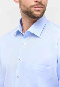 Camicia azzurra chiara con colletto aperto, chiusura a bottoni e una singola tasca sul petto. Il tessuto ha una texture liscia e leggermente lucida.