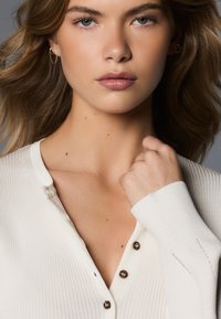 Jeune femme aux cheveux châtain clair portant un haut blanc côtelé boutonné, tenant le col près de son cou, expression neutre.