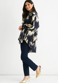 Navy and cream patterned blouse with a loose fit, long sleeves, and a curved hem, paired with dark jeans and pointed gold heels.