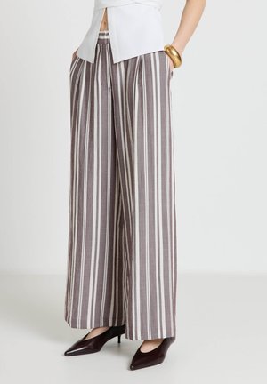 Donna che indossa pantaloni a vita alta a gamba larga con strisce verticali marroni e bianche, abbinati a tacchi scuri a punta e una maglietta bianca.