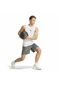 Una pelota medicinal negra sostenida por una persona que lleva una camiseta sin mangas blanca y pantalones cortos grises, en una pose de ejercicio. Zapatillas deportivas blancas visibles.