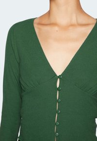 Grünes, geripptes Langarmoberteil mit tiefem V-Ausschnitt und sechs passenden Knöpfen auf der Vorderseite. Mit subtilen Raffung am Bust.