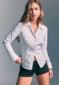 Chemise blanche boutonnée en coton ferme, avec manches longues, col classique et coupe légèrement cintrée, associée à un short noir.
