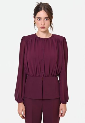 Donna con capelli castani che indossa orecchini dorati, una camicetta bordeaux a maniche lunghe con bottoni e pantaloni abbinati, in piedi su sfondo bianco.