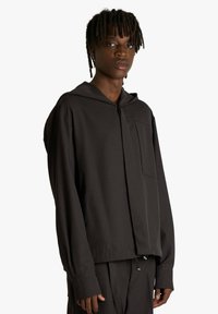 Giacca nera con cappuccio in tessuto morbido, dotata di tasca frontale e maniche lunghe con vestibilità rilassata. Linee pulite e design minimale.