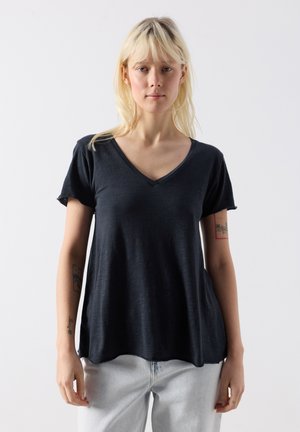 Blondīne sieviete, valkājot vaļīgu melnu V-veida kakla T-kreklu un gaiši zilas džinsas, stāv pret vienkāršu balto fonu.