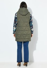 Olivově zelená prošívaná vesta s kapucí, zdobená diamantovým vzorem, nošená přes košili s dlouhým rukávem v mnoha barvách s květinovým potiskem a modré zvonové džíny.