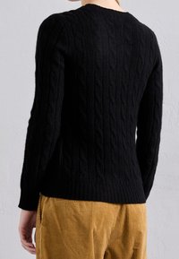 Schwarzer Zopfmuster-Pullover mit rundem Ausschnitt, gerippten Manschetten und Saum. Der Stoff wirkt dick und strukturiert. Über goldenen Cordhosen getragen.