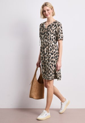 Młoda kobieta uśmiecha się, ma na sobie sukienkę do kolan w panterkę, białe trampki i trzyma dużą brązową zamszową torebkę.