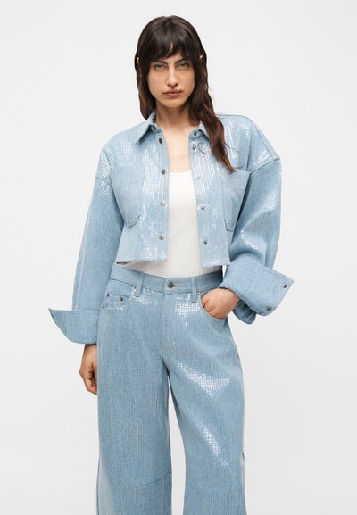 Γυναίκα φοράει cropped τζιν μπουφάν σε ανοιχτό μπλε με παγιέτες και τζιν παντελόνι καμπάνα στο ίδιο σχέδιο, με λευκό τοπ από μέσα.