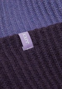 Tessuto a maglia viola con una texture a coste caratterizzato da una finitura morbida e pelosa. Un'etichetta di tessuto di colore viola chiaro è visibile sulla sezione più scura.