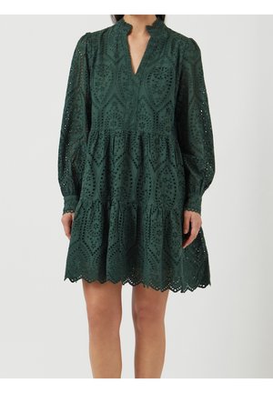 Robe verte foncée à manches longues en broderie anglaise avec jupe à volants, ourlet festonné et décolleté en V discret, portée par une personne debout sur un fond uni.