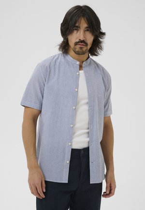 Homme aux cheveux mi-longs foncés et barbe, portant une chemise à manches courtes rayée bleu et blanc sur un débardeur blanc.