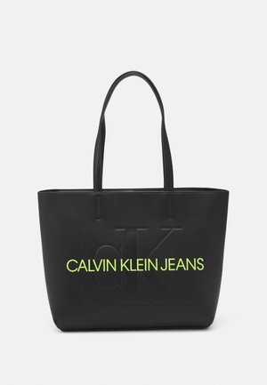 Černá taška s embosovaným logem, se neonově zeleným nápisem "CALVIN KLEIN JEANS". Hladký povrch, dvojité uchy a hranatý tvar.