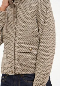 Veste à motifs marron et beige avec fermeture éclair, col à revers et poche avant ornée d'un bouton doré.