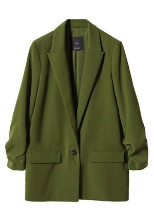 Personne portant un blazer vert olive, un t-shirt blanc, un jean bleu taille haute et des chaussures noires pointues, debout devant un fond uni.