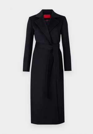 Μαύρο παλτό τύπου wrap με μακριά μανίκια, φαρδιές πέτσετες και ζώνη στο ίδιο χρώμα· έχει λεία υφή και φτάνει έως το γόνατο. Εμφανής κόκκινη εσωτερική φόδρα.