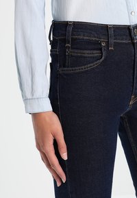 Jeanbyxor i mörkblått med orange sömmar, med framfickor och en närbild av en ljusblå skjortärm som visar knappdetaljer.