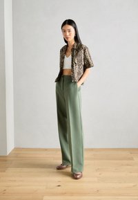 Chemise à manches courtes imprimée serpent marron, crop top blanc, pantalon large vert, et chaussures marron, sur fond gris clair.