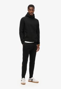 Zwarte hoodie met een hoge kraag en kangoeroe-pocket, gecombineerd met zwarte joggers. Witte sneakers met zwarte accenten. Eenvoudig, minimalistisch ontwerp.