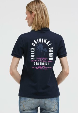 Frau mit kurzem blondem Haar, die ein marineblaues Poloshirt mit der Aufschrift „SOCCX Original Brand Sea Breeze“ und einem Palmenmotiv auf dem Rücken trägt.