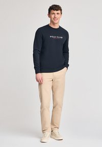 Marinblå sweatshirt med "POLO CLUB"-logotyp, beige byxor och ljusa sneakers. Slät tygkvalitet, rundad halsringning, ribbad mudd och nederkant.