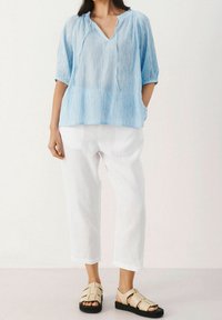 Helles, strukturiertes, hellblaues Pulli mit V-Ausschnitt und kurzen Ärmeln, kombiniert mit weißen, lockeren Hosen und beigen, sandalettenartigen Schuhen.
