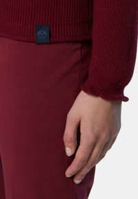 Maglione a coste bordeaux con polsini ondulati, abbinato a pantaloni in tessuto bordeaux coordinati. Include un piccolo etichetta blu del marchio in vita.