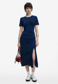 Mörkblå klänning med korta ärmar, rynkdetaljer, glipor på sidan och abstrakta svarta tryck; kombinerad med vita strumpor och svarta och vita skor, hållande en röd väska.