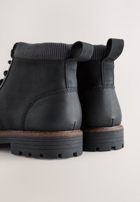 Ghete negre din piele pentru gleznă, cu talpă de cauciuc, toc suprapus și manșetă striată; prevăzute cu bretele pentru tras și cusături pe toc pentru durabilitate sporită.