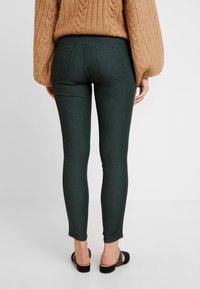 Jeans skinny verde oscuro de cintura alta, con bolsillos traseros y una textura suave, combinados con un suéter de punto beige y chanclas negras.