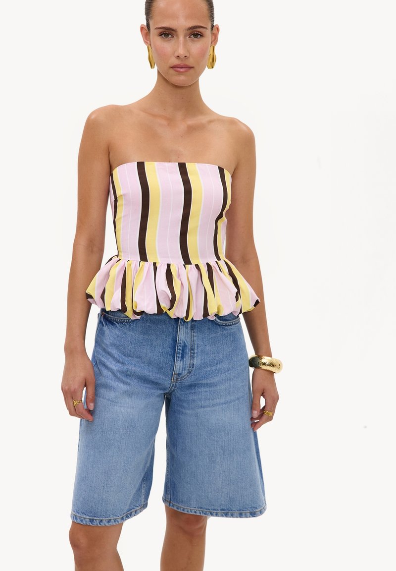 Femme portant un haut bustier à volants avec des rayures verticales marron, rose et jaune, un short en jean bleu, des boucles d'oreilles en or, un bracelet et des bagues.