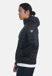Giacca a vento nera con design trapuntato orizzontale, cappuccio e patch con logo sulla spalla sinistra. Tessuto liscio e lucido con una vestibilità aderente.
