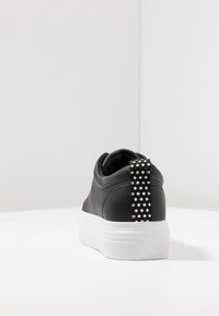 Černé tenisky s bílou podešví, vybavené texturovaným černým páskem na patě zdobeným bílými puntíky. Hladký materiál s zaobleným tvarem.