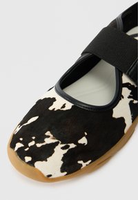 Sandal estilo Mary Jane com padrão de pele de vaca preto e branco, tira elástica preta e sola de borracha bege sobre fundo branco.