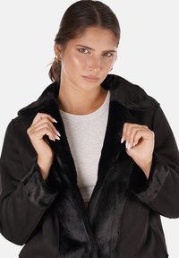 Giovane donna dai capelli castani tiene il colletto in finta pelliccia nera del suo cappotto, indossando un top grigio chiaro sotto, su uno sfondo semplice.