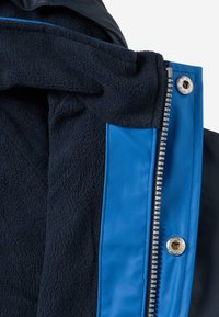 Gros plan d'une veste bleue montrant une doublure en polaire noire, une fermeture éclair argentée et un bouton-pression métallique sur la coque extérieure bleue.