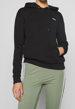 Henkilö, jolla on musta FILA-huppari, jossa on kiristysnauhat, ja vihreät leggingsit, joissa valkoiset sivuraidat, toinen käsi hupparin taskussa.