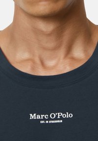 Close-up of a person's neck and upper chest wearing a dark blue shirt with "Marc O’Polo EST. IN STOCKHOLM" printed in white.