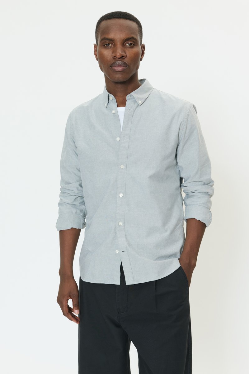 Camisa gris clara abotonada con cuello, mangas remangadas y bolsillo frontal, combinada con pantalones negros de corte holgado. Textura de tela suave.