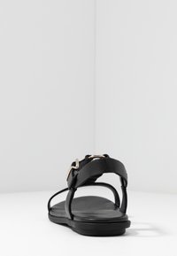 Svart lädersandal med platt sula, öppen tå och justerbar rem med metallisk detalj för stängning. Enkel och minimalistisk design.