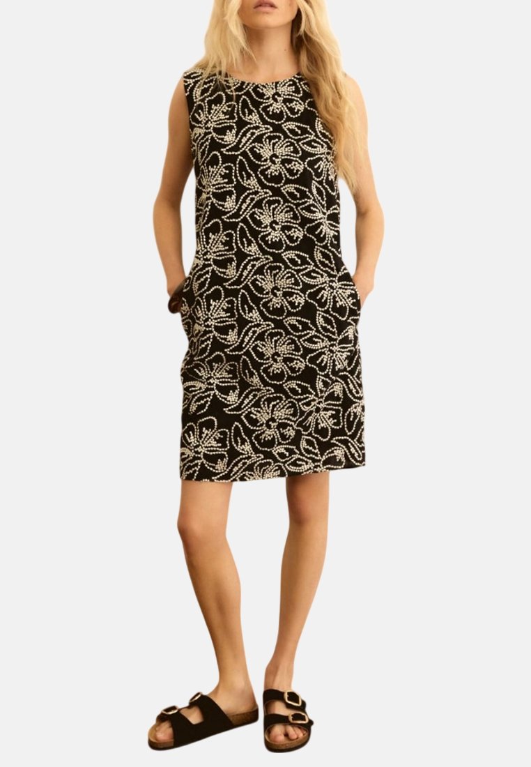 Donna che indossa un abito nero senza maniche con motivi floreali bianchi e sandali neri slip-on con fibbie, in piedi con le mani in tasca.