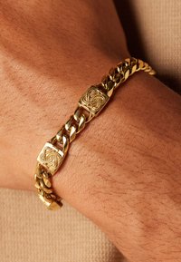 Gouden metalen schakelarmband met vergrendelde schakels en twee vierkante accenten met ingewikkelde ontwerpen. Soepele textuur op de huid.