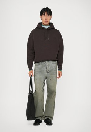 Bruine hoodie met verhoogde tekst; lichtgroene spijkerbroek met versleten accenten; zwarte totebag; zwarte schoenen; neutrale achtergrond.