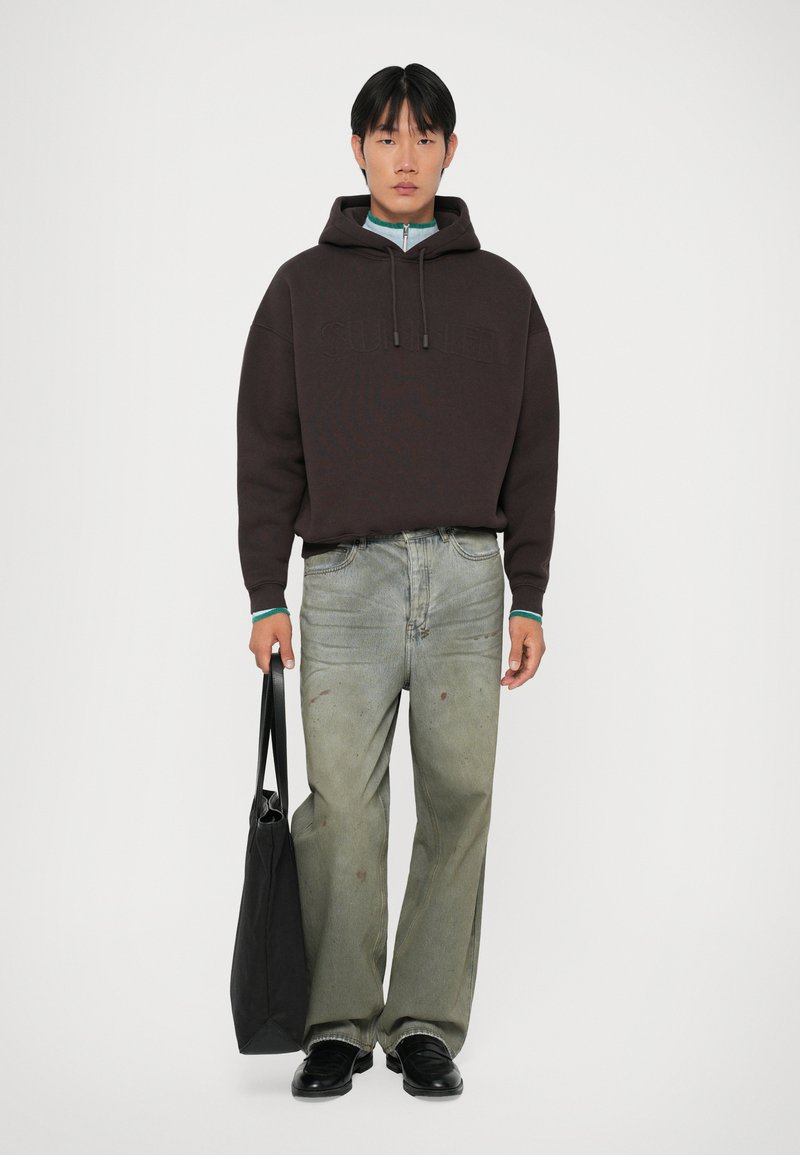 Felpa con cappuccio marrone con testo in rilievo; jeans verde chiaro con effetti scoloriti; borsa tote nera; scarpe nere; sfondo neutro.