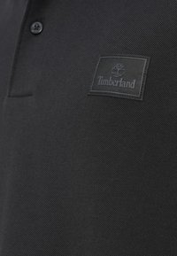 Μαύρο πόλο μπλουζάκι από υφασμάτινη υφή με ραμμένο ένθετο λογότυπο Timberland στην αριστερή πλευρά. Δύο κουμπιά κοντά στον γιακά.